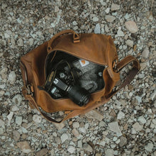 Cargar imagen en el visor de la galería, The Dagny Weekender | Large Leather Duffle Bag on gravel with camera inside, showcasing spaciousness and organization for travelers.