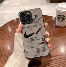 Cargar imagen en el visor de la galería, Luxury leather sneaker phone case featuring a sleek design and branding, held over a wooden table next to a coffee cup.