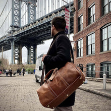 Cargar imagen en el visor de la galería, Man carrying The Dagny Weekender | Large Leather Duffle Bag near iconic bridge and brick buildings, showcasing style and durability.