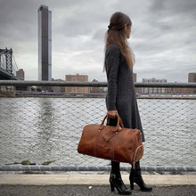 Cargar imagen en el visor de la galería, Woman standing with The Dagny Weekender | Large Leather Duffle Bag by a river in an urban setting