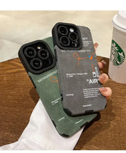 Cargar imagen en el visor de la galería, Two Luxury Leather Sneaker Phone Cases with sleek design and suede labels held over a wooden table next to a Starbucks cup.