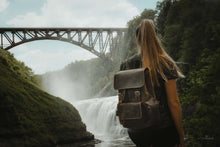 Charger l'image dans la galerie, Woman exploring with The Hagen Backpack | Vintage Leather Backpack near a scenic waterfall and bridge.