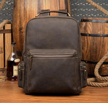 Load image into Gallery viewer, The Langley Backpack | Genuine Vintage Leather Backpack displayed in a rustic setting with wooden barrels and rope in the background.
