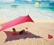Charger l'image dans la galerie, Man relaxing under eZthings UV sun shade beach shelter on sandy shore, sunny day, red canopy with sandbag anchors, ocean view.