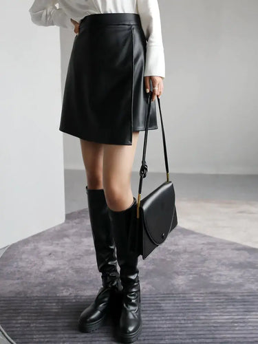 Woman wearing a black high waist PU leather A-line split short skirt, paired with boots and a handbag.