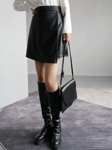 Woman wearing a high waist black PU leather A-line split short skirt with knee-high boots and holding a black handbag.
