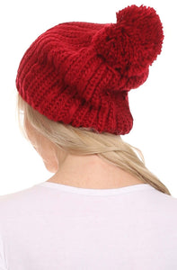 Red cable knit beanie with large pom pom, worn by model, showcasing slouchy fit and wide design for versatile wear.