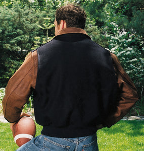 Man wearing REED varsity jacket with leather sleeves, standing outdoors holding a football, showcasing American-made quality.